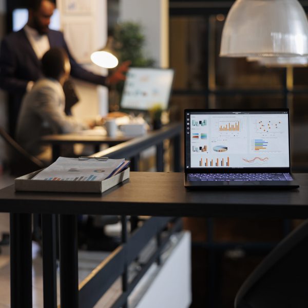 laptop-computer-with-financial-graphs-screen-standing-table-startup-office-coworkers-discussing-company-strategy-background-employees-working-late-night-startup-office-scaled.jpg